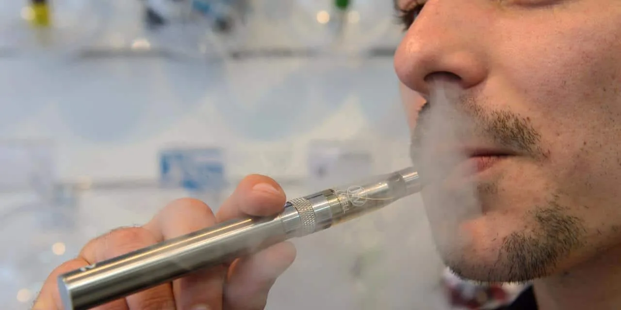 Salarié tenant une cigarette électronique devant un bureau en open space, avec un panneau d’interdiction visible en arrière-plan