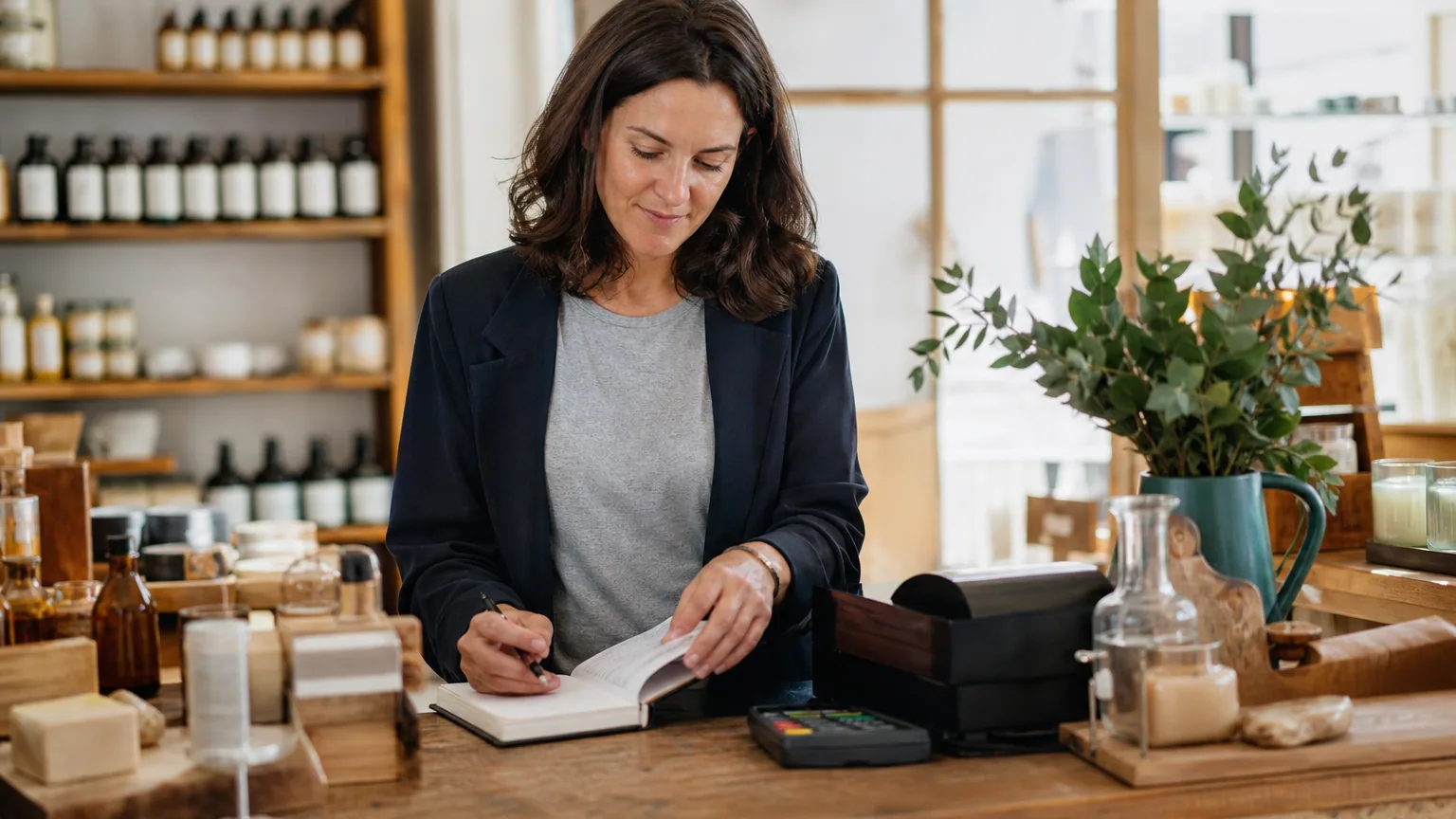 Commerçante derrière son comptoir vérifiant un carnet de ventes à côté d'étagères de produits et d'un terminal de paiement